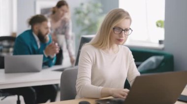 Ofiste bilgisayarını kullanan çekici bir kadın ve arka planda meslektaşları var. Günlük kıyafet uzmanı istatistiksel verileri grafiklerde analiz ediyor..