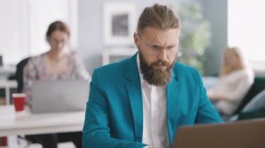 Handsome male worker browsing necessary information on digital laptop in office room. Concentrated bearded man preparing presentation for meeting with female colleagues sitting behind.