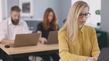 Businesswoman working on laptop in busy office with her colleagues on background. Focused entrepreneur browsing internet while planning ideas in startup agency.
