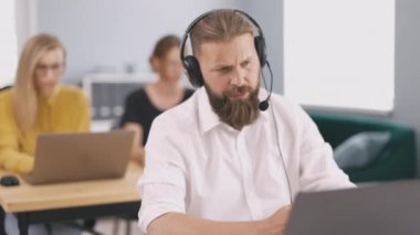 Caucasian male employee in headset having video call on modern laptop. Focused man introducing company possibilities to potential customer. Office workers on background.