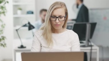 Mature blonde businesswoman wearing eyeglasses typing on laptop sitting in light crowded office