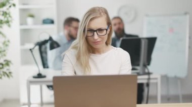 Smiling satisfied businesswoman in eyeglasses typing on laptop. Light office indoors shot