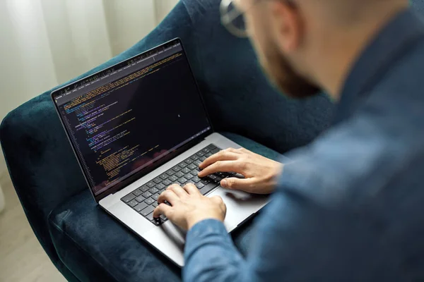 Young man in denim jacket coding on laptop while sitting on blue couch, remote networking concept