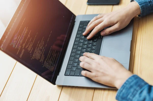 Man coding on laptop, screen full of code, crop view of desk with computer and hands