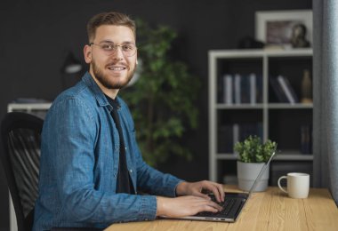 Laptop 'a kod yazan yakışıklı bir gencin portresi, koyu renk ofiste kot gömlek giyen bir programcı.