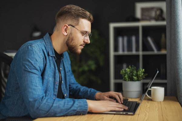 Young handsome bearded man writing code on laptop, programmer wearing denim shirt and eyeglasses in dark office, it concept