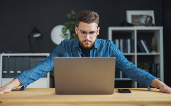 Confident young man in eyewear focused on laptop screen, bearded coder thoughts