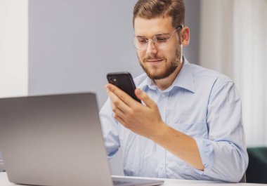 Sakallı adam akıllı telefon uygulaması kullanıyor. Evde dizüstü bilgisayarı var. Teknoloji konsepti.