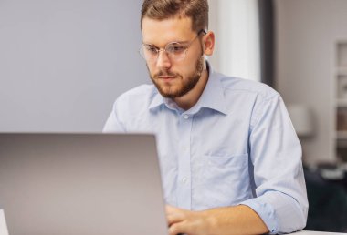 Genç odaklı sakallı iş adamı dizüstü bilgisayarla çalışıyor, projeye odaklanmış.