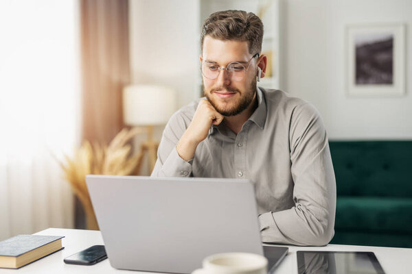 Happy young coder working with laptop from home, handsome man coding using gadgets and computer