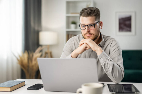 Thoughtful handsome man coding from home at laptop, remote work concept