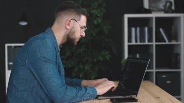 Genç sakallı kodlayıcı dizüstü bilgisayar kullanıyor, karanlık ofis odasında kodlama yapıyor, uygulama yazılımı yaratıyor.