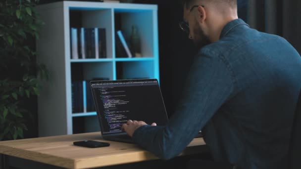デニムシャツの髭の男ノートパソコン上でコードを書く 暗いオフィスで若いプログラマーコーディングソフトウェア — ストック動画