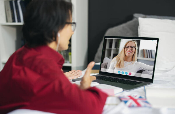 Вид с плеча зрелой женщины, использующей ноутбук для видеочата с подругой, темноволосой дамой, имеющей видеокамеру в спальне. Концепция связи людей