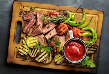 Dilimlenmiş orta boy biftek, ızgara sebze ve ketçap sosu, ahşap kesme tahtası, yakın plan koyu görünüm.