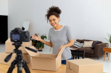 Afrikalı Amerikalı kadın blogger vlogger freelancer takipçileri için video içeriği kaydetmek için kamera kullanarak ürünlerin ve malların genel bir değerlendirmesini yapıyor. Kamerada kutuları açan genç bir kadın.