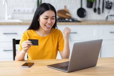 Güzel, heyecanlı Asyalı kadın portresi online alışveriş yapıyor, dizüstü bilgisayar kullanıyor. Gülümseyen mutlu kadın elinde kredi kartıyla dijital ekrana bakıyor.