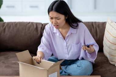 Üzgün Asyalı genç bir kadın evdeki kanepede otururken internetten yanlış ürün teslim aldı, online alışveriş, teslimat konsepti.