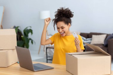 Güzel bir afro-amerikan kadının portresi online alışveriş yapıyor, dizüstü bilgisayar kullanıyor. Gülümseyen mutlu kadın elinde kredi kartıyla dijital ekrana bakıyor.