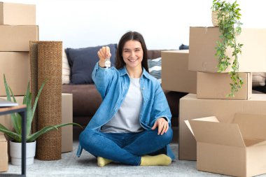 Yeni bir eve taşınmak, kiralık konutlar. Yeni evindeki karton kutularla yerde oturan ve elinde anahtarlar olan mutlu, genç beyaz bir kadın kameraya bakarak gülümsüyor.