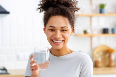Healthy lifestyle concept. Beautiful young Afro American woman holding a glass of clean fresh water standing at home in the kitchen, looks at the camera and smiles friendly