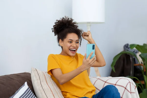 Excited young African American woman using mobile phone shopping online sitting at home on the couch. Big sales concept