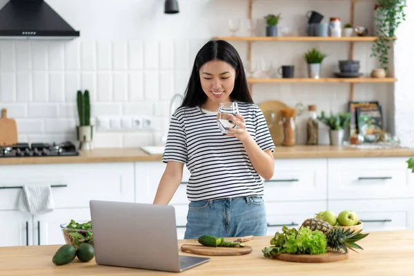 Asian young woman vlogger live broadcast cooking healthy food standing at home in the kitchen looking at laptop screen, smiling friendly