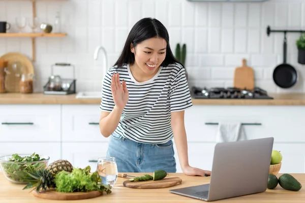 Asian young woman vlogger live broadcast cooking healthy food standing at home in the kitchen waving at the laptop, smiling friendly