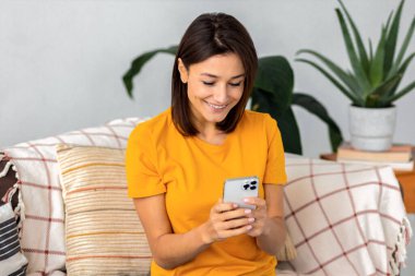 Beautiful young joyful woman using mobile phone and smiling sitting at home on the couch