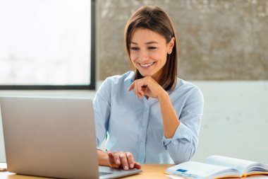 Başarılı bir girişimci kadın, modern bir ofiste oturup bilgisayar ekranına bakarken bilgisayarını kullanır ve dostça gülümser.