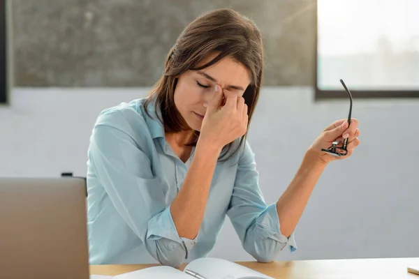 Overworked young woman office worker holds glasses in her hands, massages the bridge of her nose, sitting at her workplace in the office