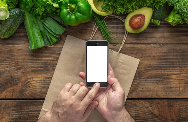 Order food online. Man holds mobile phone with blank screen on wooden background with fresh green vegetables top view