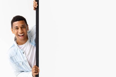 Young African American man peeking out from empty white board, looks at the camera and smiles. Copy space. Place for your advertising, mock-up