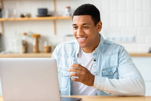 Young male student or freelancer uses laptop for online education or work, drinks fresh water. Happy African American man working from home, studying, distance learning