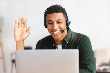 African american young man customer support call center operator or receptionist sitting at the workplace in a modern office consulting a client, uses a headset, smiles friendly
