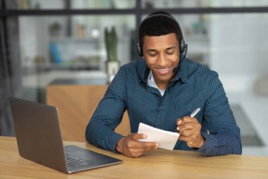 Portrait of confident African American young man. Successful student or office worker in headset sitting at workplace and working using laptop, taking notes in a notebook, smiling friendly