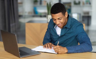 Portrait of confident African American young man. Successful student or office worker sitting at workplace and working using laptop, smiling friendly