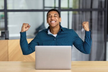 Portrait of excited young African American man celebrating success while sitting with laptop in the workplace in the office. Happy businessperson male having good news, he receive payment