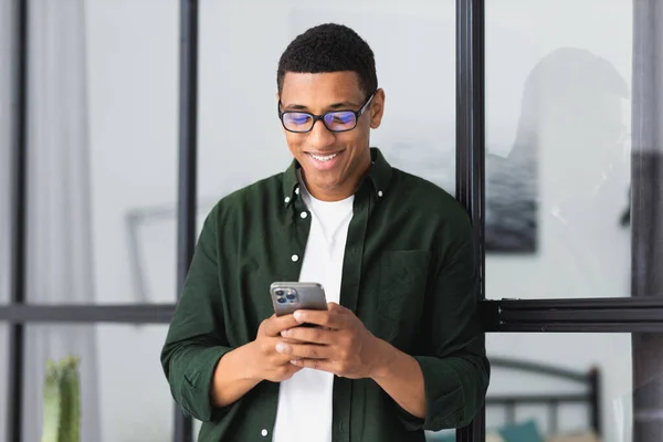 Young smiling African American man relaxing using modile phone, working in modern office. Male entrepreneur looking at screen typing message with smartphone. Happy guy playing online games