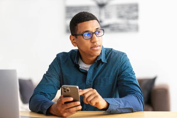 Focused young businessman holding a mobile phone, looking away and thinking about something Portrait of African American businessman in glasses sitting at the workplace in the office with a smartphone