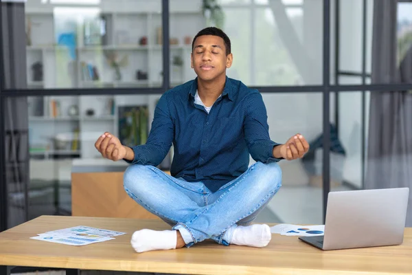 Calm African American young man sitting on the workplace in the office and meditating taking break avoiding stressful job