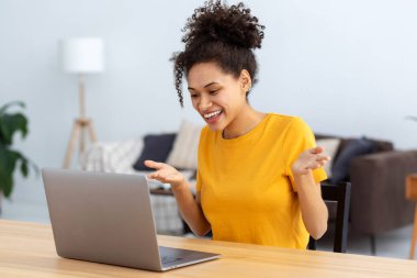 Laptop bilgisayarı kullanan mutlu Afrikalı Amerikalı kız öğrencinin portresi. Ev ofisinde oturmuş, dil öğrenirken görüntülü konuşma yapıyor. Günlük giysiler içinde serbest çalışan, uzaktan çalışan, bir şeyleri tartışan biri.