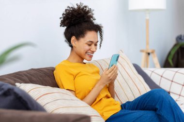 Genç Afro-Amerikalı kadın cep telefonu kullanıyor, internette sohbet ediyor, evinde kanepede oturuyor. Afro kadın akıllı telefon ekranına bakar ve dostça gülümser.