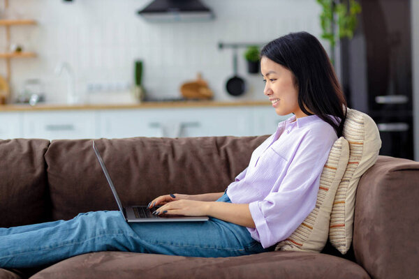 Счастливая азиатка с помощью ноутбука сидит на уютном диване дома в чате в социальной сети или ищет что-то в Интернете