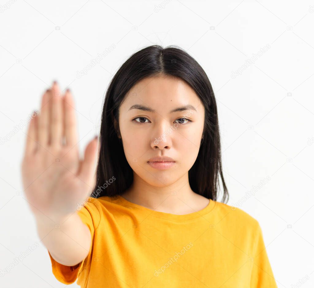 Portrait of young serious Asian woman showing stop gesture and looking at camera on a white background