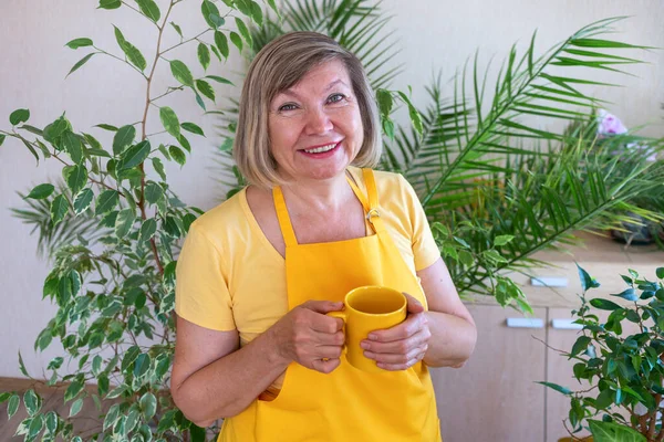 Старша жінка п'є фрілансер чаю в жовтому фартуху в зеленому будинку, відпочиває в гармонії з рослинами навколо. Домашній сад, фрілансер — стокове фото