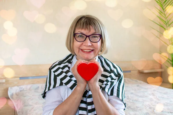 Senior woman Valentines Day with a heart in her hands on the background of her bedroom at home. Senior Citizens Day.