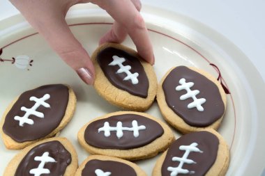 Futbol şeklinde kurabiyeler. Elin tabaktan kurabiye alıyor. Amerikan spor anlayışı. Super Bowl parti kurabiyeleri.