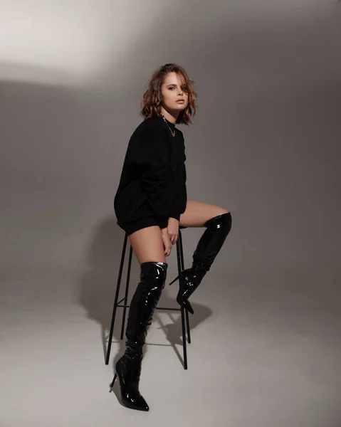 白いスタジオを背にポーズをとる美人 ブラックのスウェットシャツとブラックのブーツを着たモデル バーの椅子に座ってる女性 ファッションポートレート — ストック写真