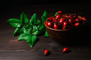 Stüdyo resmi, koyu ahşap arka planda yeşil dallı kırmızı gül kalçalı.